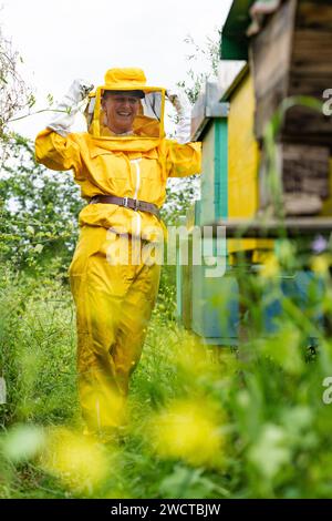 Apiculteur adulte positif en costume de protection jaune avec voile et gants debout et souriant dans le rucher Banque D'Images