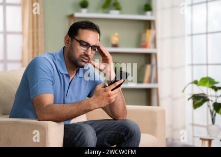 Homme indien choqué a eu de mauvaises nouvelles tout en utilisant le téléphone portable sur le canapé à la maison - concept de détresse financière, annulation de prêt et avenir incertain Banque D'Images