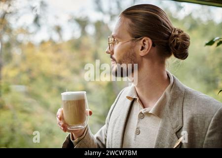 bel homme attrayant avec la barbe et les cheveux recueillis dans un costume élégant buvant son café chaud Banque D'Images