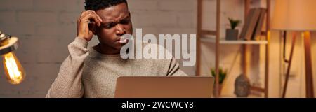 freelance afro-américaine sérieuse travaillant à l'ordinateur portable dans le bureau à domicile la nuit, bannière horizontale Banque D'Images