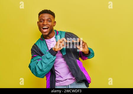 homme afro-américain débordé de joie dans une veste lumineuse tenant des lunettes de soleil et dansant sur fond jaune Banque D'Images