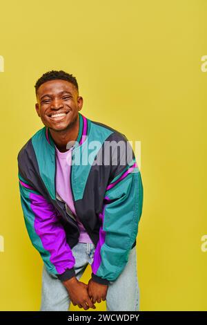 homme afro-américain à la mode débordé de joie dans le brise-vent lumineux souriant et posant sur fond jaune Banque D'Images