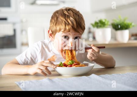 Un mignon petit garçon mange des spaghetti bolognaise pour le déjeuner dans la cuisine à la maison et est couvert de ketchup. Banque D'Images