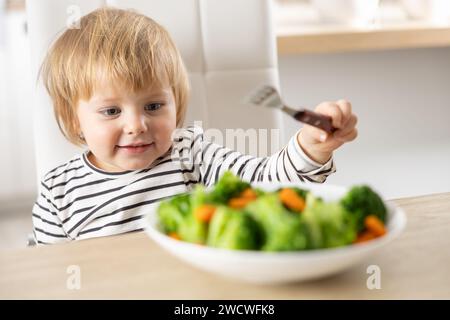 Mignonne petite fille mange des légumes de brocoli et de carottes avec une fourchette. Banque D'Images