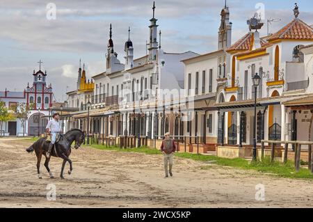 Espagne, Andalousie, El Rocío, cavalier sur un cheval noir aux côtés d'un vieil homme vêtu d'un béret dans une rue sablonneuse bordée d'hermandades, les maisons des confréries catholiques Banque D'Images