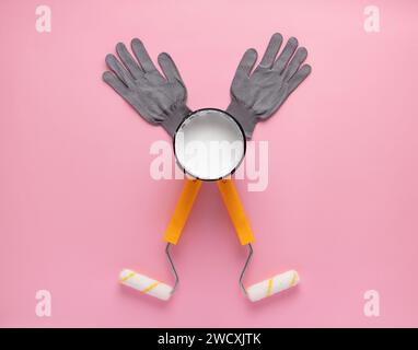 Composition créative de rouleaux à peinture, boîte ouverte de peinture blanche et gants de travail sur fond rose. Visualisation de l'homme heureux avec les mains levées doi Banque D'Images