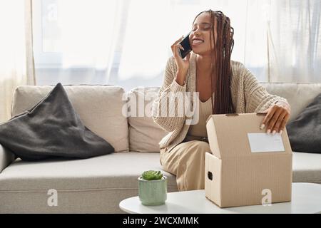 joyeuse femme afro-américaine attrayante dans des vêtements de maison confortables parlant par téléphone avec la main sur le colis Banque D'Images