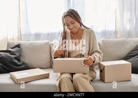 belle femme afro-américaine joyeuse en homewear assise sur le canapé et regardant la boîte en carton Banque D'Images