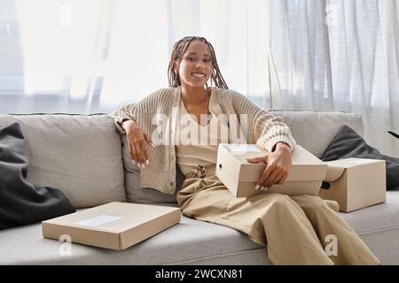 joyeuse jeune femme afro-américaine en tenue confortable assise sur un canapé avec des boîtes et souriant à la caméra Banque D'Images