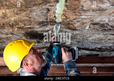 un travailleur en salopette et un casque de protection tient une perceuse visseuse à batterie dans sa main et visse le cadre dans le bois, qu'il utilise t Banque D'Images