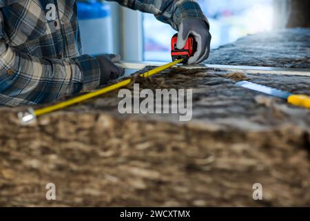 un travailleur en salopettes et gants mesure la longueur de la laine de verre avec un mètre. Banque D'Images