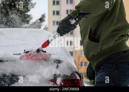 un gars en veste, chapeau et gants balaie la voiture après une tempête de neige avec un petit balai rouge. Banque D'Images