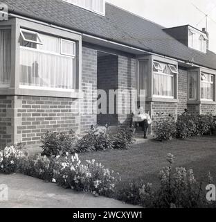 Années 1960, historique, un gentleman assis dans un transat lisant un livre à l'extérieur sur la marche avant d'un nouveau bungalow chalet, Angleterre, Royaume-Uni. Banque D'Images