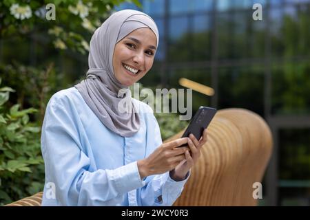 Une femme musulmane souriante en hijab exploite un smartphone à l'extérieur avec un immeuble de bureaux en toile de fond, exsudant confiance et professionnalisme. Banque D'Images