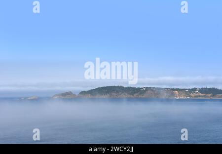 Une vue lointaine à travers la baie montre un quai de chargement à sec, un quai de dolly, à Port Orford, Oregon. Le brouillard est bas sur la surface de l'océan mais le ciel bleu est au-dessus. Banque D'Images