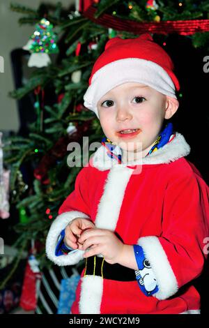 Jeune garçon, vêtu d'un costume de Père Noël, prend une pause de l'ouverture des cadeaux pour manger du chocolat le matin de Noël. Banque D'Images