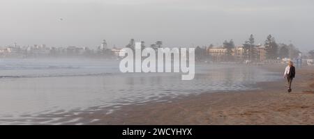 Femme marche le long d'une plage de sable fin au coucher du soleil avec une tour dans la médina derrière, à Essaouira, Maroc, 17 janvier 2024 Banque D'Images