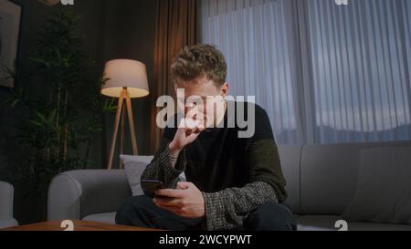 Beau jeune homme blond assis sur le canapé à la maison tapant un message texte par smartphone. Personne masculine se relaxant à la maison. Communiquer avec la famille et frie Banque D'Images