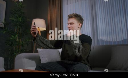 Bel homme blond assis sur le canapé ayant la conversation d'appel vidéo avec le téléphone intelligent. Partagez des nouvelles, parlez à votre famille, profitez de réunions virtuelles de chat en ligne Banque D'Images