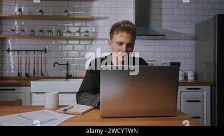 Jeune homme d'affaires beau assis à la maison dans la cuisine regardant son ordinateur portable, vidéoconférence, parler. Travail à domicile, freelance, télétravail, Banque D'Images