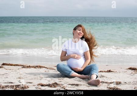 Heureuse femme blanche enceinte assise sur une plage près de l'océan. Bonne grossesse Banque D'Images