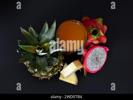 Un grand verre d'un mélange de jus de fruits exotiques sur fond noir, à côté d'un ananas mûr et de morceaux de pitaya. Vue de dessus, pose à plat. Banque D'Images