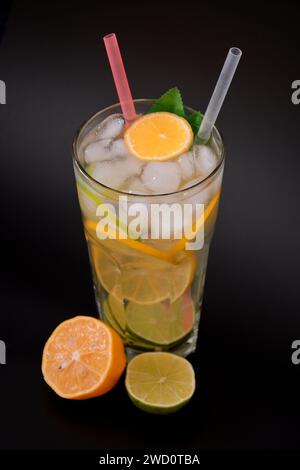 Limonade rafraîchissante dans un grand verre à facettes avec de la glace et des morceaux d'agrumes sur fond noir, à côté d'une demi-orange mûre et de citron vert. Arrangeurs verticaux Banque D'Images