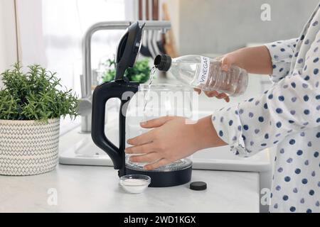 Femme versant du vinaigre de bouteille dans une bouilloire électrique dans la cuisine, gros plan Banque D'Images