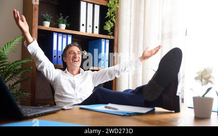 Homme heureux à bras ouverts assis dans son bureau avec ses pieds sur la table en bois avec des documents et des étagères avec classeurs derrière Banque D'Images