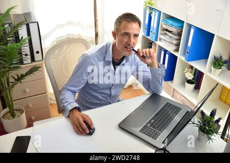 Entrepreneur indépendant pensatif mordant un stylo dans son bureau à domicile assis sur une chaise dans un bureau blanc avec des étagères pleines de dossiers et une fenêtre dans le Banque D'Images