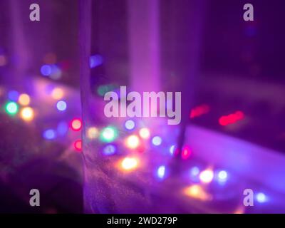 Lumières de fées à cordes floues multicolores brillant sur le rebord d'une fenêtre de nuit sombre derrière le rideau de filet, se reflétant dans le verre. Banque D'Images