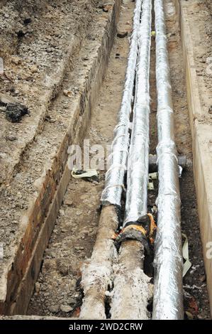 Installation de conduites d'eau chaude souterraines avec isolation, conduites de chauffage urbain dans des tranchées près d'un bâtiment résidentiel. Banque D'Images