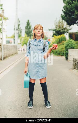 Portrait en plein air d'une petite écolière drôle, portant une robe formelle et un sac à dos, tenant une boîte à lunch et une pomme rouge. Concept de retour à l'école, film look ton Banque D'Images