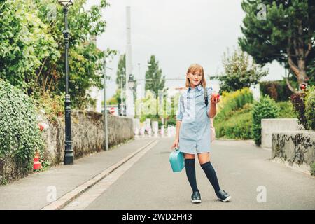 Portrait en plein air d'une petite écolière drôle, portant une robe formelle et un sac à dos, tenant une boîte à lunch et une pomme rouge. Concept de retour à l'école, film look ton Banque D'Images