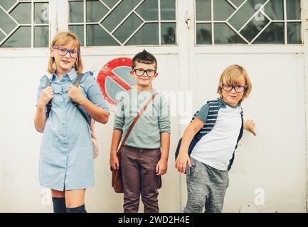 Groupe de trois enfants drôles portant des sacs à dos marchant de retour à l'école. Fille et garçons portant des lunettes posant à l'extérieur Banque D'Images