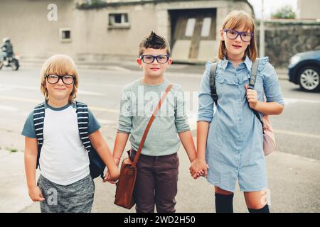 Groupe de trois enfants drôles portant des sacs à dos marchant de retour à l'école. Fille et garçons portant des lunettes posant à l'extérieur à côté de la route Banque D'Images