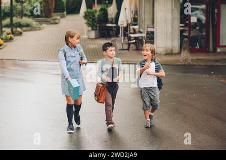 Groupe de 3 écoliers drôles qui retournent à l'école ensemble, portant des sacs à dos, tenant une boîte à lunch et une planche à roulettes Banque D'Images