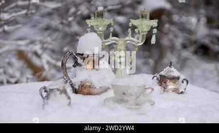 Un pot à café argenté, un pot à lait, un pot à sucre dans la neige. Une tasse avec de la glace. Un bougeoir. Table jouée pour la reine de glace. Banque D'Images