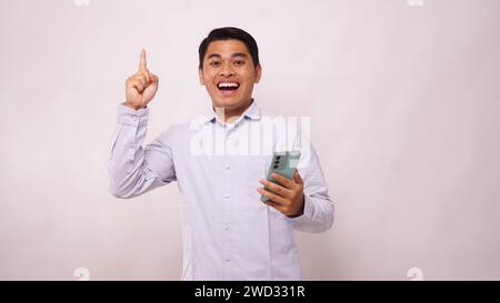 Homme asiatique poing serré montrant l'excitation en regardant son téléphone sur fond blanc Banque D'Images