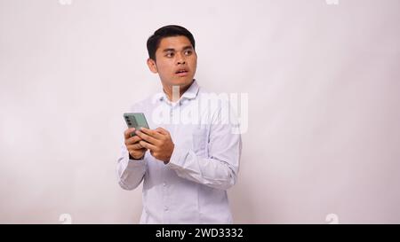 Homme asiatique gratter sa tête tout en tenant le téléphone portable et montrant l'expression confuse du visage sur fond blanc Banque D'Images