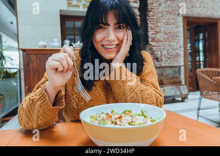 jeune femme assise penchée de la table avec une main sur son visage surpris heureux souriant avec fourchette et bol de salade, regardant la caméra Banque D'Images