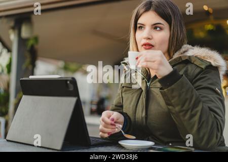 Une jeune femme courbée et concentrée dans une veste verte profite d'une tasse de café chaud pendant qu'elle travaille sur sa tablette, incarnant la flexibilité du télétravail. Banque D'Images