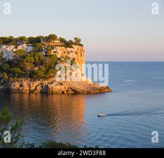 Cala Galdana, Minorque, Îles Baléares, Espagne. Vue sur la mer calme aux falaises de calcaire escarpées, coucher de soleil, bateau de retour au port. Banque D'Images