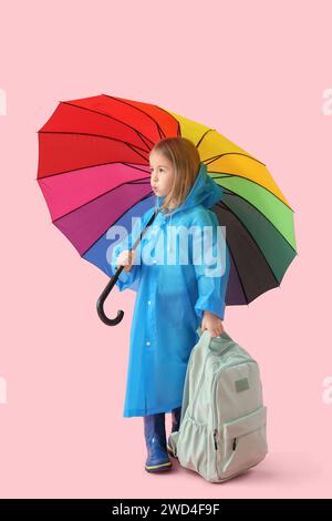 Mignonne petite écolière en imperméable avec sac à dos et parapluie marchant sur fond rose Banque D'Images