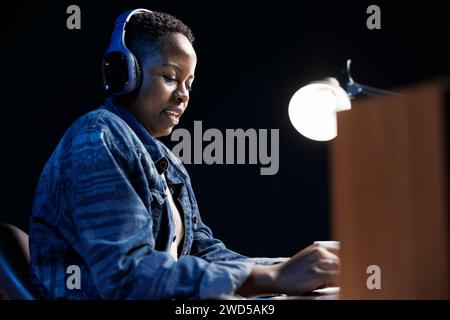 Jeune femme afro-américaine utilisant un ordinateur portable dans l'appartement, naviguant sur Internet et écoutant de la musique avec des écouteurs sans fil. Femme freelance avec micro-casque travaillant à distance, vérifiant les e-mails. Banque D'Images