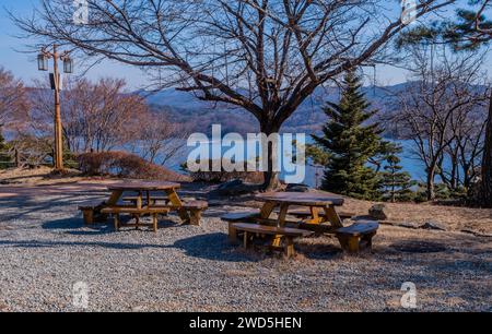 Deux tables de pique-nique rondes en bois devant un arbre sans feuilles avec lac et montagnes en arrière-plan, Corée du Sud, Corée du Sud, Corée du Sud Banque D'Images