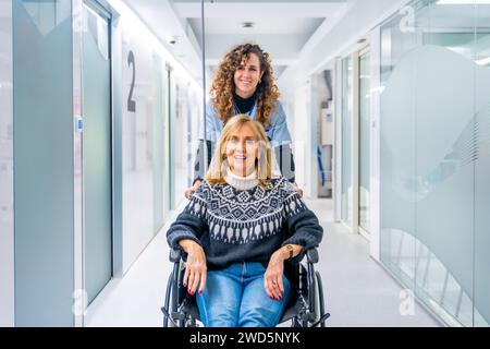 Infirmière heureuse transportant une patiente adulte en fauteuil roulant dans le couloir hospitalier Banque D'Images