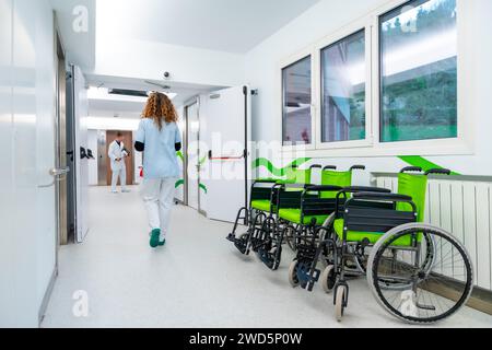 Vue arrière d'une infirmière marchant le long d'un couloir d'un hôpital avec des fauteuils roulants stationnés Banque D'Images