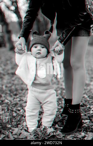 Petit garçon apprend à faire les premiers pas en tenant les mains de maman en automne dans le parc. Photo noir et blanc avec effet de glitch 3D. Banque D'Images