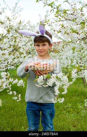 Chasse aux œufs de Pâques. Mignon garçon préscolaire portant des oreilles de lapin tenant le panier plein d'oeufs de Pâques dans les mains à la chasse aux oeufs de Pâques dans le jardin. Portrait contre le dos Banque D'Images